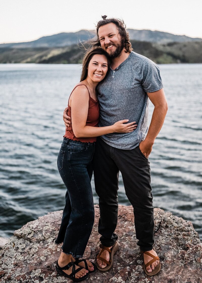 Portrait of the husband and wife team behind Alpine Vows, Colorado elopement photographer and videographer duo, standing on a rocky lakeshore with mountain views in the background