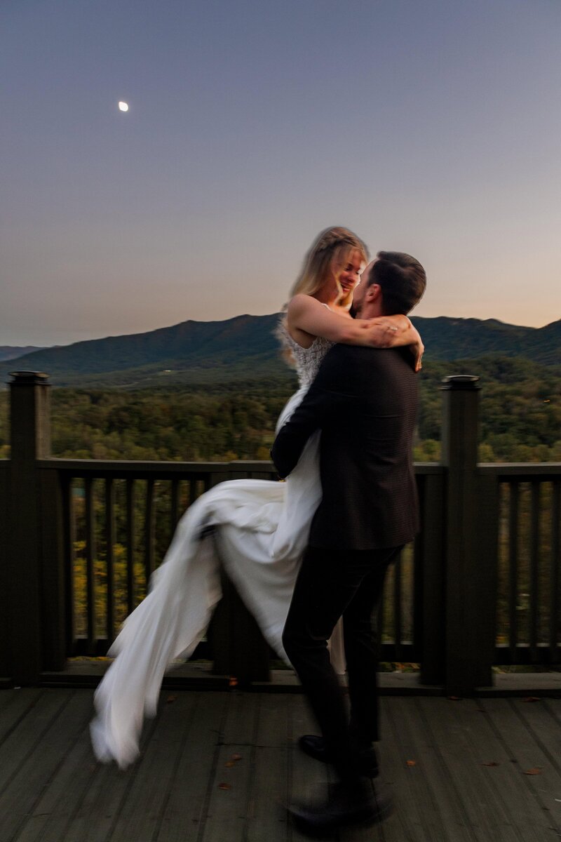 A groom is lifting and spinning his bride around after getting married. They are standing in front of a vast mountain range as the moon is rising behind them. They are looking lovingly at each other while smiling and laughing. 