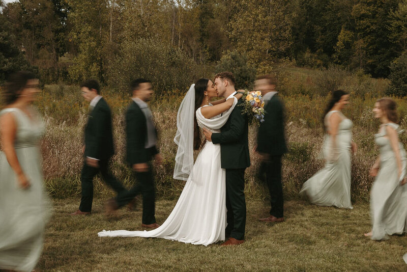 Bride and groom kissing as the bridal party walks behind them, creating a blur of motion during their Michigan wedding