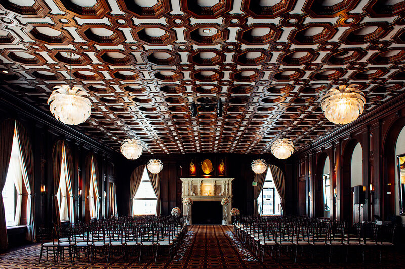 Professional San Francisco wedding photography showcasing Julia Morgan Ballroom ceremony setup with black chairs, ornate hand-carved coffered ceiling, crystal chandeliers, dark wood paneling, and grand fireplace creating luxurious historic atmosphere