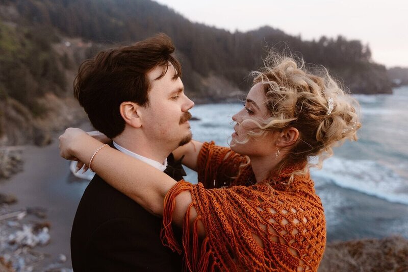 A couple stands close by the river in the Redwoods, with forested hills in the background. The bride wears a burnt orange shawl and has her arms around the groom’s neck during their elopement.