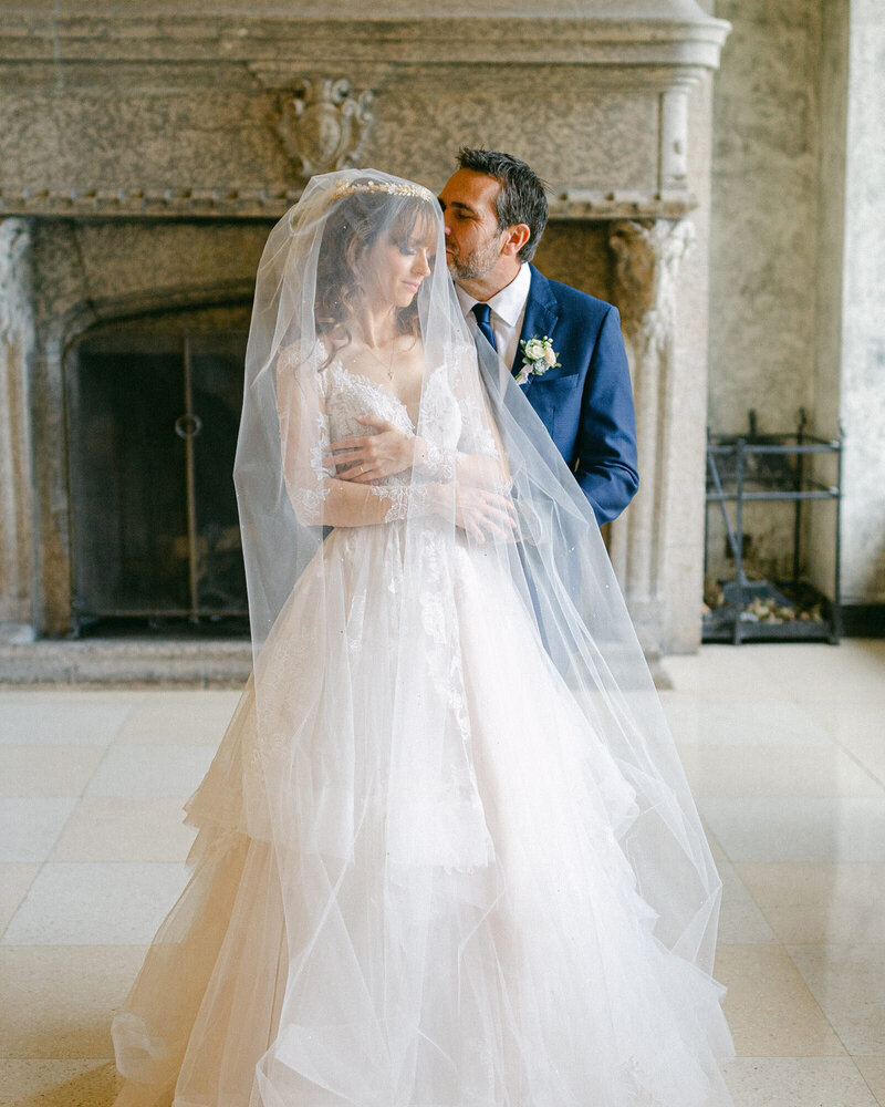 Bride and groom in the Riverview hallway at the Fairmont Banff Springs Hotel with the groom gently kissing the bride through her veil and a fireplace in the background captured as a luxury wedding moment