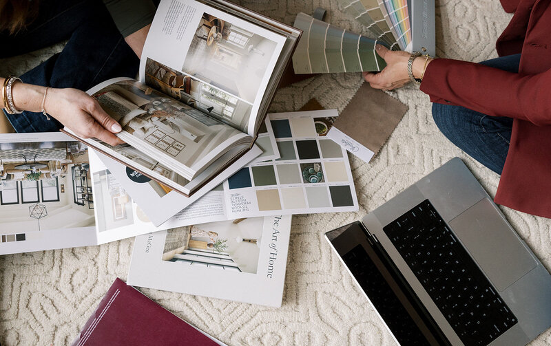 Two people sitting on a textured rug are browsing interior design books and examining color swatches. A laptop is open nearby, and various magazines and books are spread around, showcasing home decor ideas and inspiration.