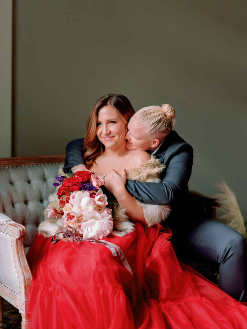 A couple sits indoors on a couch, one kissing the other's neck.