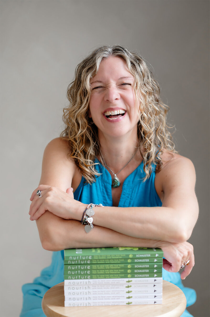 Heidi Schauster stands in front of a white wall, next to a vibrant potted plant, smiling warmly at the camera.