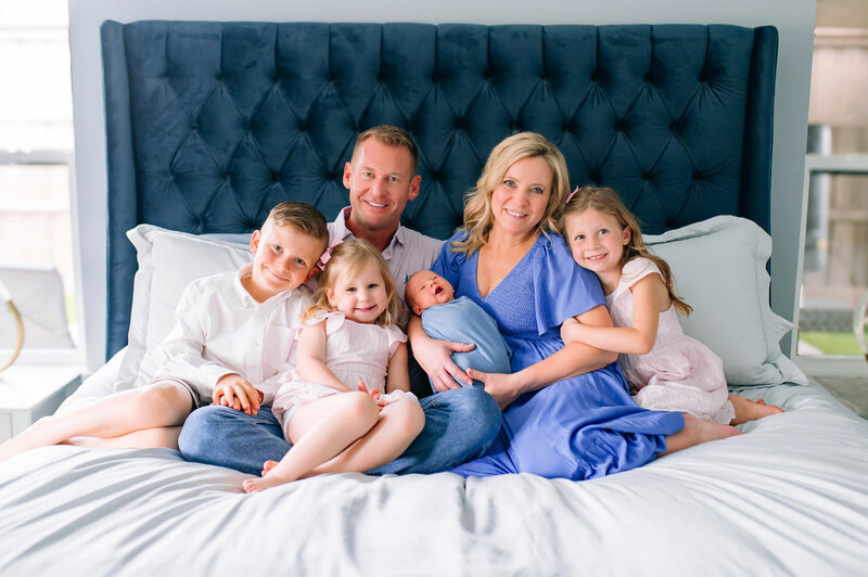 Parents sitting on a bed with their four young children during an in-home family and newborn session in Allen, Texas, captured by Jennifer L. Kirk Photography.