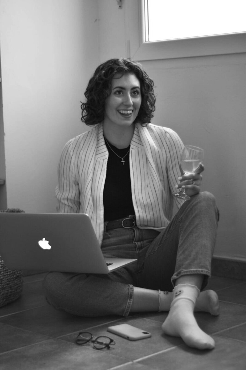Emily sits on the floor, smiling and holding a glass of wine and laptop, she has a striped blazer, jeans, and socks with smiley faces on them