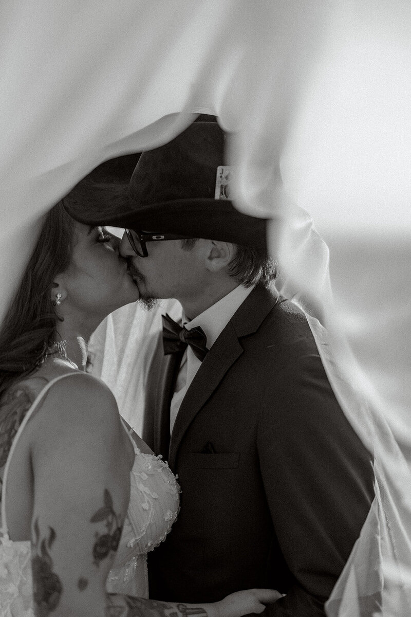 Captured in a dreamy moment, the couple shares an intimate kiss beneath the flowing veil, bringing romance and emotion to their California wedding. The groom, in his sharp suit and signature cowboy hat, perfectly complements the bride’s tattooed elegance in her lace gown. This black-and-white shot beautifully frames their connection and unique love story against the soft breeze of the California coast. A timeless memory from a truly unforgettable day.