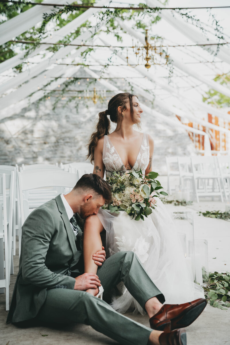 bride sitting in a chair holding a bridal bouquet while groom kisses her leg
