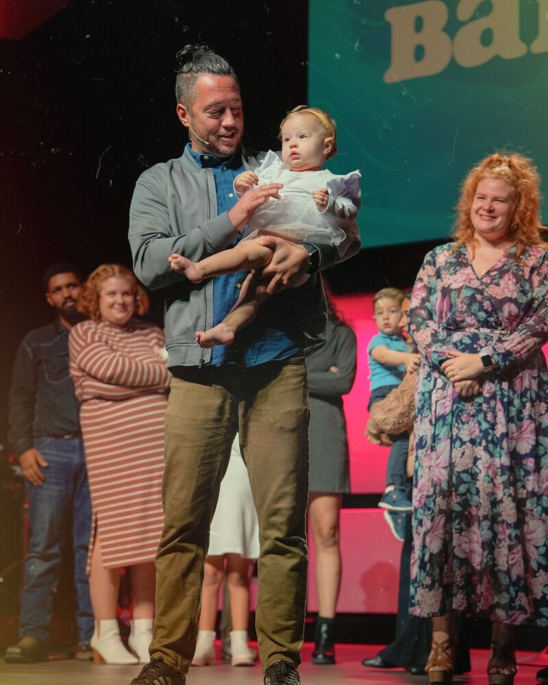 Pastor Matt holding a baby on stage while onlooker smile.