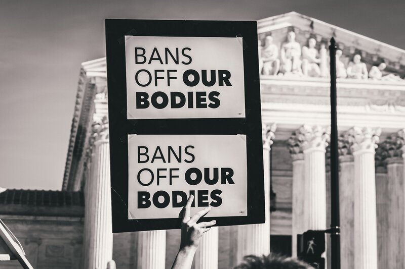A sign that reads, "Bans Off Our Bodies," is being held in the air in front of a government building in the background. The image is black and white.