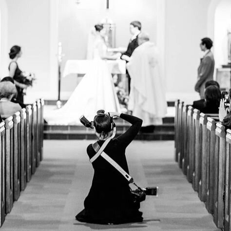 Black and white photo of a wedding ceremony taken from the aisle. A photographer kneels in the centre, capturing the moment as the bride and groom stand at the altar with the officiant. Guests are seated on either side, witnessing the event.