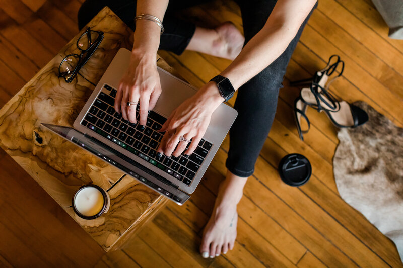 Overhead view of Allie Clark typing on a MacBook Pro at a wood coffee table, with accessories like glasses, a candle, and heels nearby — a behind-the-scenes moment from Allie Clark Designs, highlighting the polished yet approachable creative process.