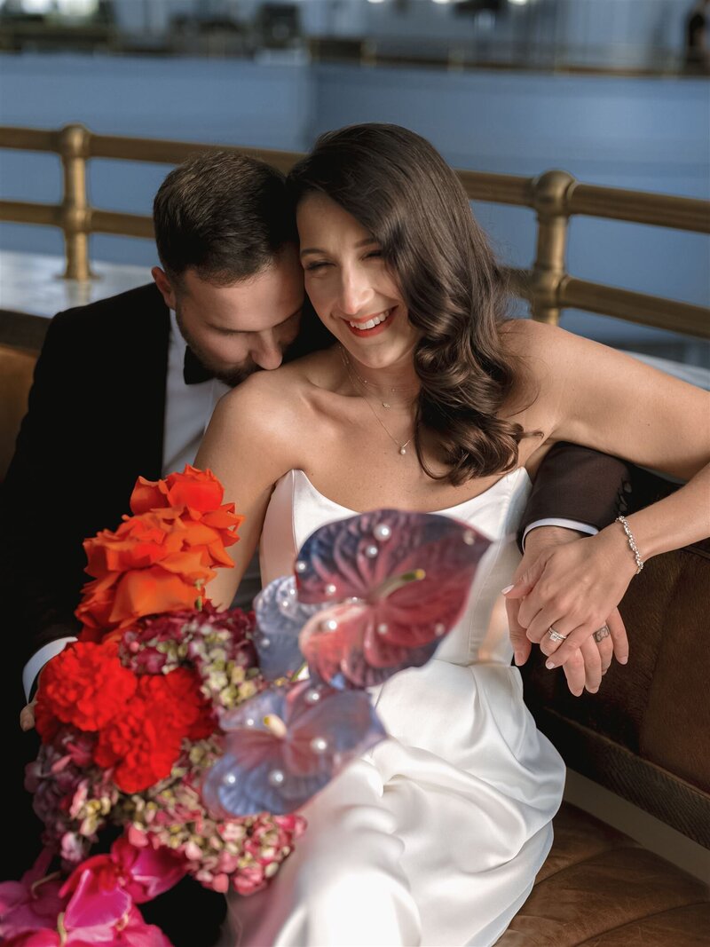 Bride and groom sitting on a leather coach on the mezzanine of the Line Hotel DC with the groom snuggling her shoulder and bride smiling and giggling holding her bouquet in her lap