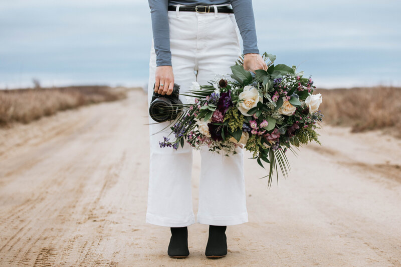A woman on a dirt road holding a camera and large bouquet of flowers wearing white pants, blue top, and black boots