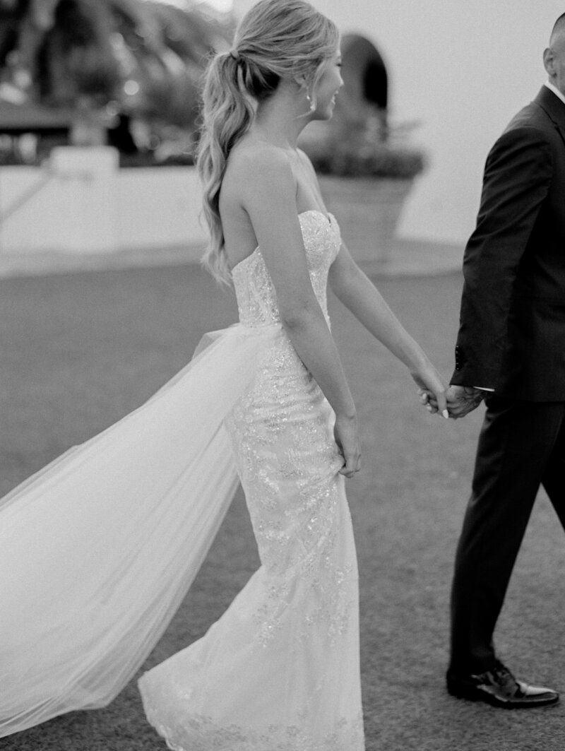 A black and white photo of a newly married bride and groom sitting at the head table, smiling. The table has a large wine through the centre of the table, they are outside.