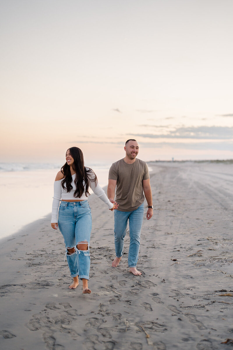 Couple walks holding hands at sunset in Cape May, New Jersey for a romantic engagement session