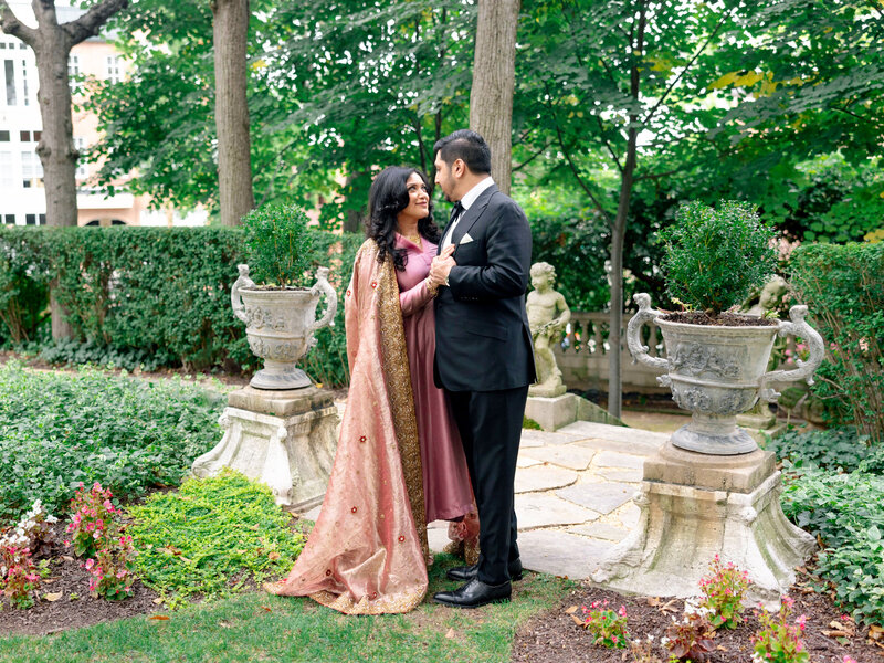 A couple stands in a lush garden, smiling at each other. The woman wears a traditional pink and gold dress, while the man is in a black suit. They are surrounded by greenery, stone planters, and a neatly manicured hedge.