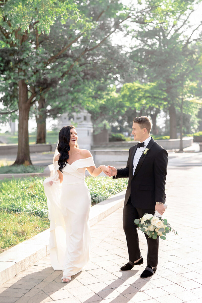 A bride in a white dress and a groom in a black tuxedo hold hands and smile at each other while walking on a sunlit path surrounded by trees. The groom holds a bouquet of white flowers.