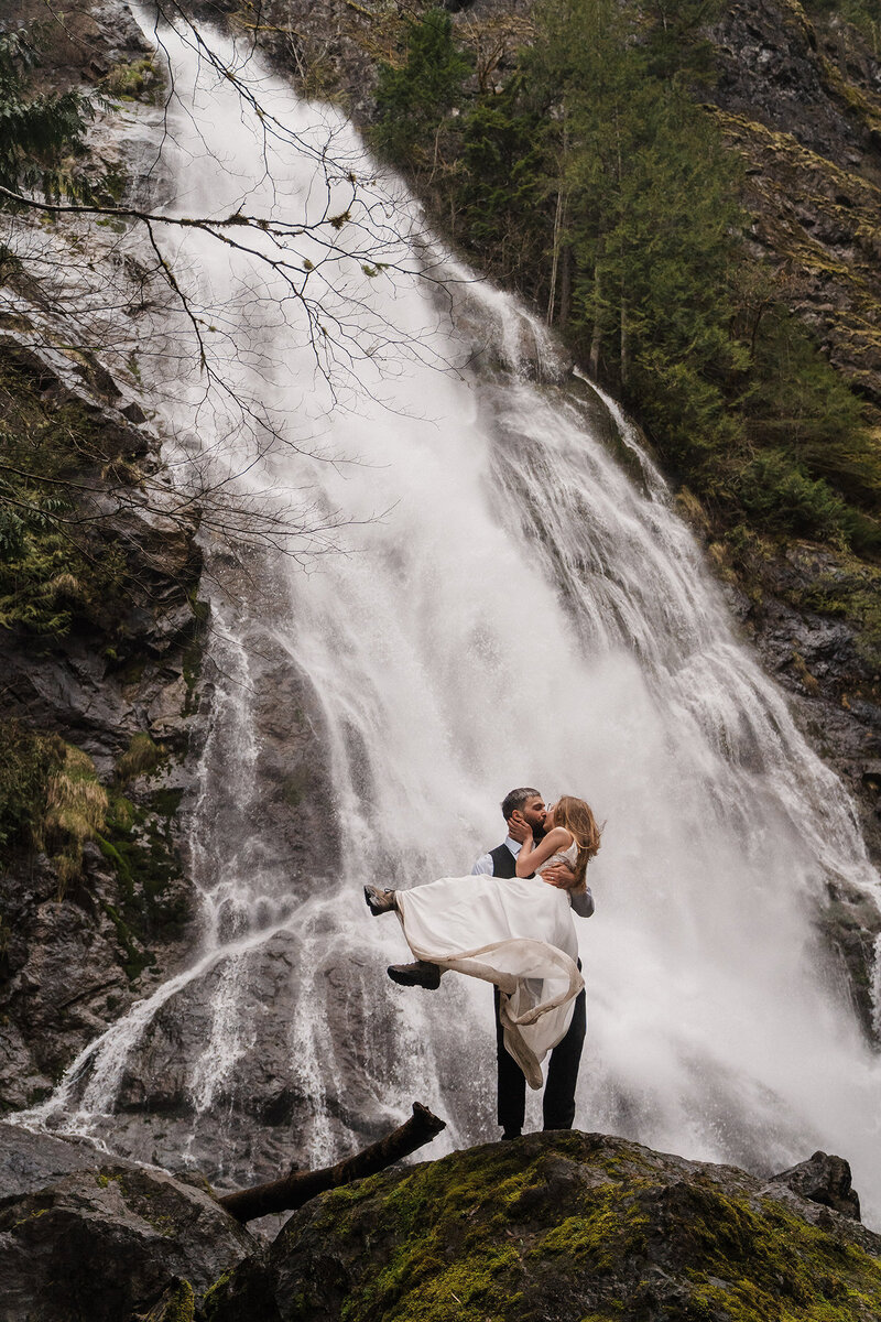 a groom lifts his bride in front of a towering waterfall near Olympic National Park