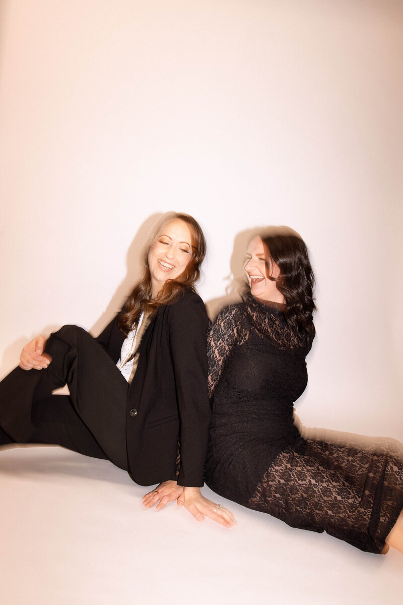 Two women sit on the floor back to back, laughing. One wears a dark suit and heels, the other woman wears a long lacy dress. The photo has a soft, slightly blurred effect and is tilted to the right. 