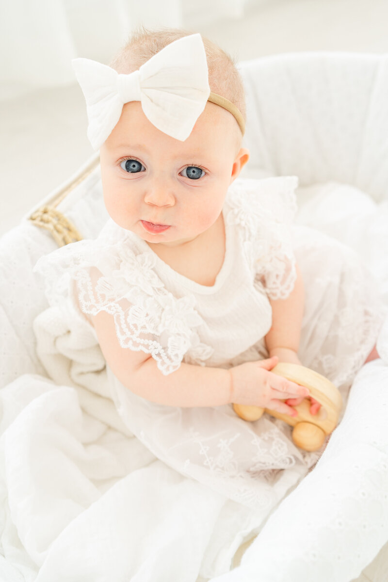 Austin, TX Family & Newborn Photography | A young baby girl sits in a moses basket in an all-white studio. She has big blue eyes and wear a big white bow in her hair.