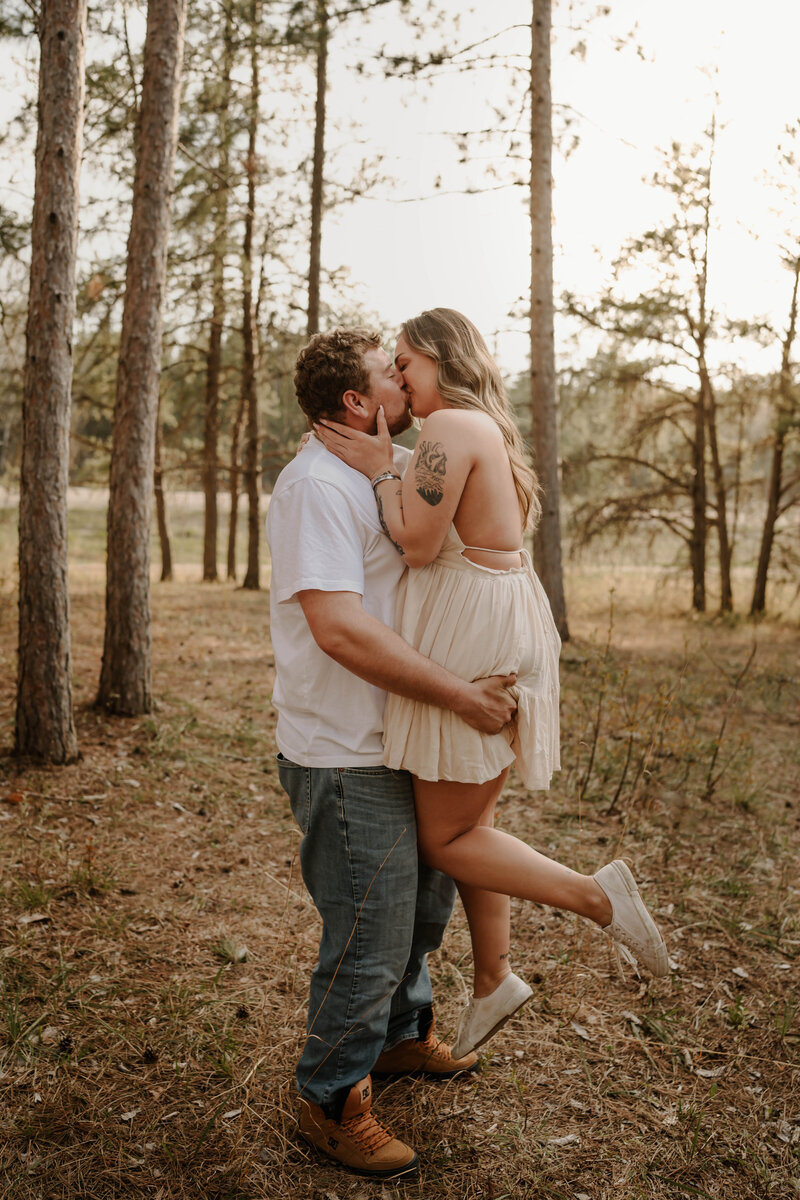 Boyfriend Lifts his girlfriend up as they lean in for a kiss surrounded by trees