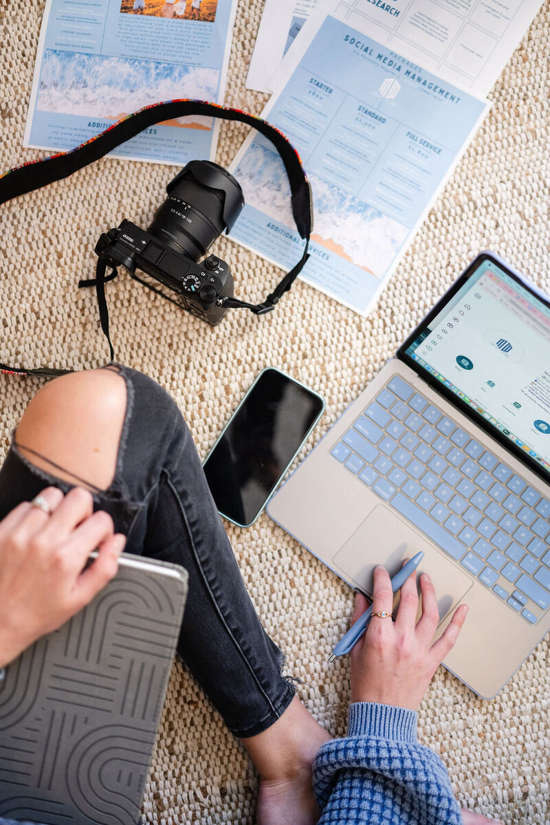 woman holds a pen while working on her laptop with camera, phone, and flyers set out on the floor