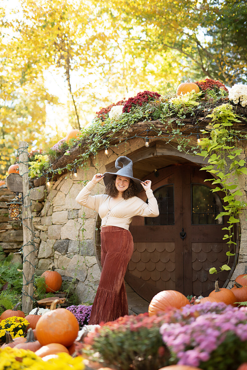 brown curly hair women poses in front of Hobbit House while holding hands on hat