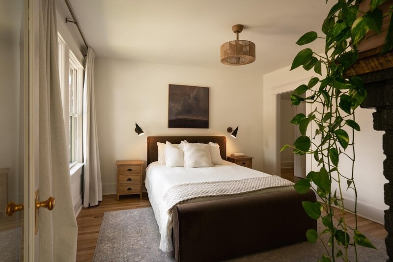 Soft-lit master bedroom with dark wood bed frame, white bedding, matching wooden nightstands, modern wall sconces, hanging green pothos plant, and rattan overhead light.