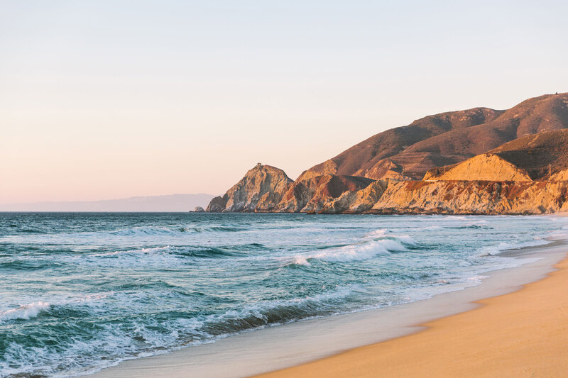 A scenic view of a beach at sunset, with gentle waves lapping on the sand. Rocky cliffs rise in the background, bathed in warm, golden light. The sky is clear, with a soft gradient of colors from blue to orange.