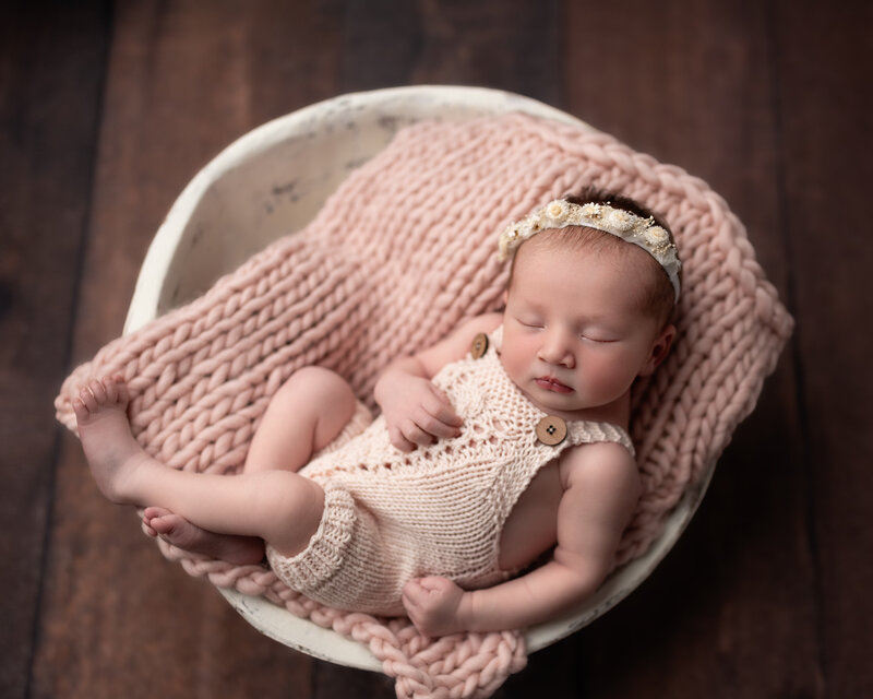 Newborn baby girl styled in a knit romper with floral headband, posed in bowl with blush textures.