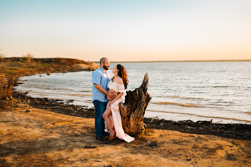 A pregnant woman and her husband stand on a beach at sunset. The mom is leaning against a tree stump and looking at her husband and smiling.
