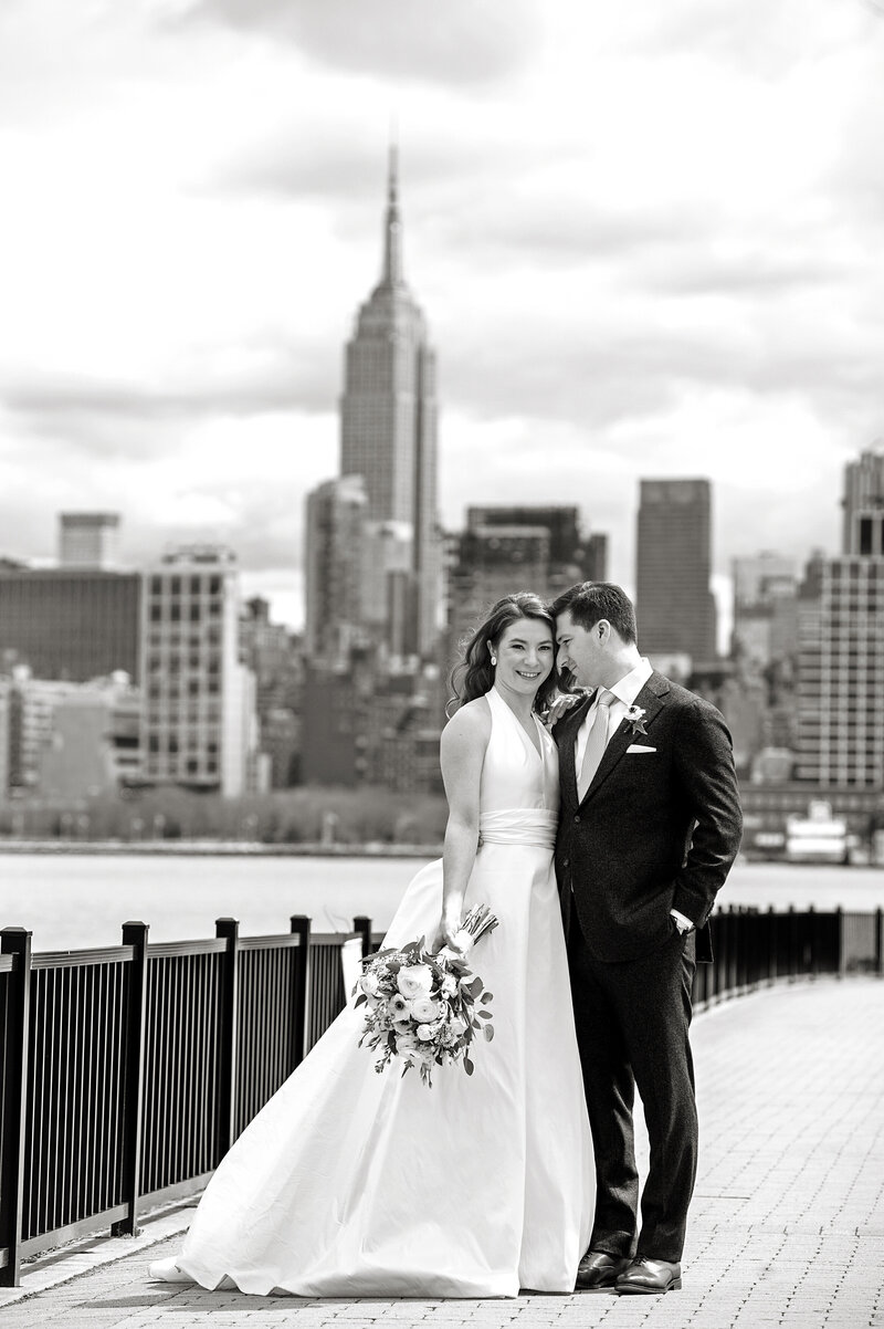 A newlywed couple standing on the edge of the water with a city skyline behind them 