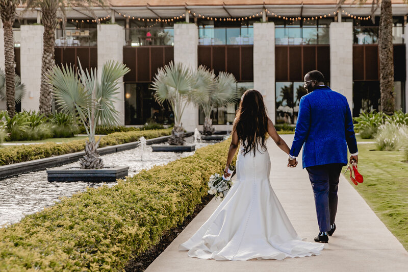 A bride and groom walk hand in hand along a landscaped pathway with water features and greenery. The bride wears a strapless white gown and holds her shoes, while the groom, dressed in a blue suit, carries the bride’s red shoes.