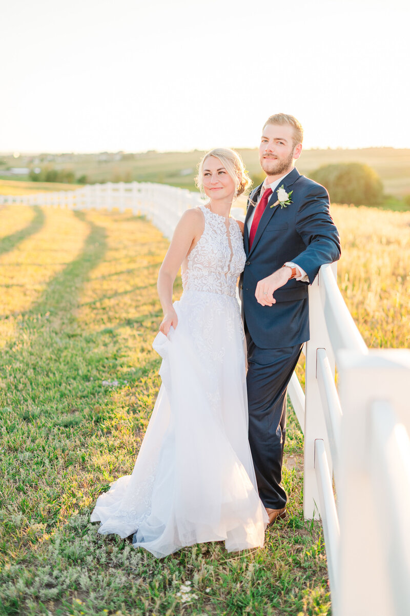 Bride and groom leaning against a white fence in a field at sunset on their wedding day.