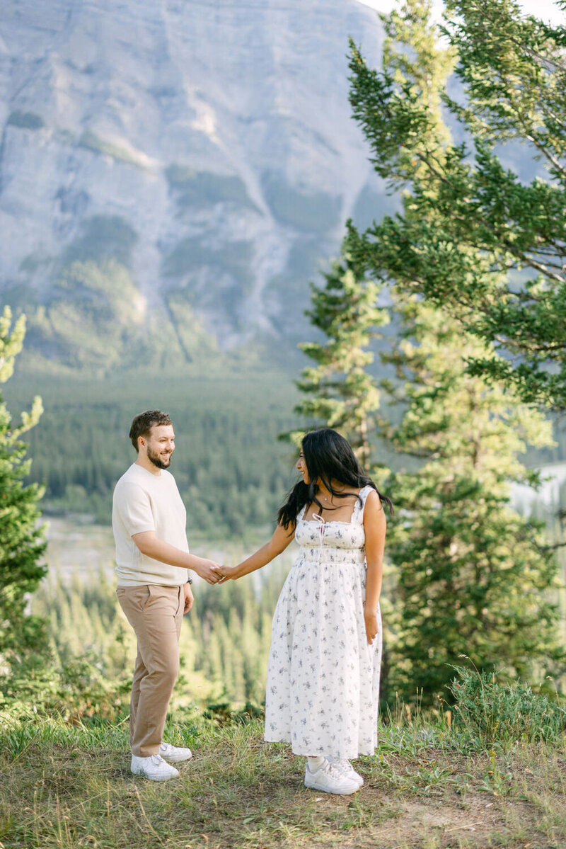 This warm image features a fiancée hugging her fiancé from behind among the snowy woods of Lake Louise. Their smiles reflect a joyful connection, capturing a tender moment of love and laughter in the serene, wintry forest setting.