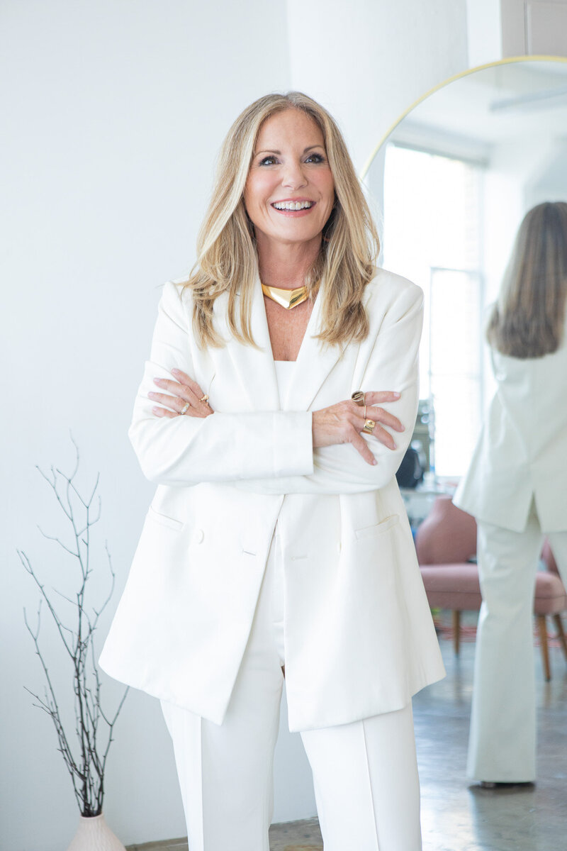 woman in a white suit smiling with arms crossed standing in a modern room with a mirror reflecting her image