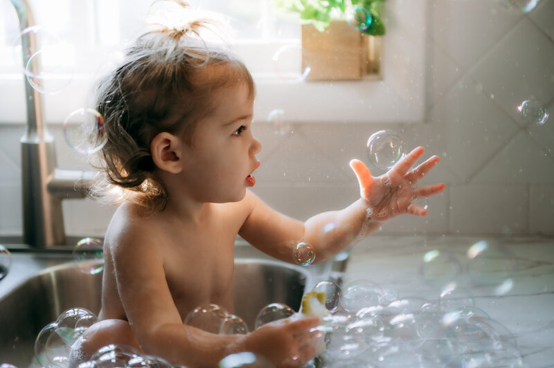 Little girl sitting in the kitchen sink surrounded by bubbles during a candid in-home session.