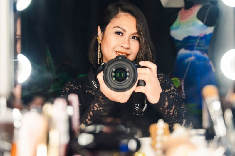 Color portrait of a female photographer in a black lace dress and gold earrings, holding a Sony camera in front of a mirror with makeup items on the table.