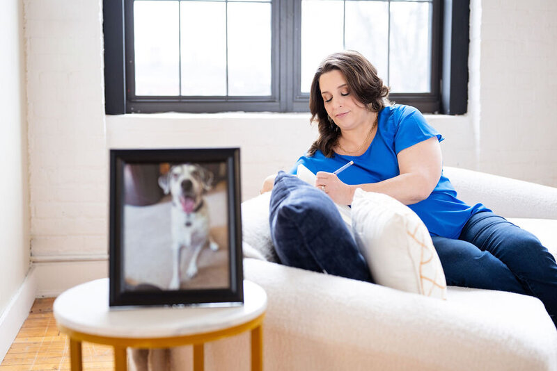 Dr. Chris Limone journaling on a couch in a bright room, with a framed photo of her dog in the foreground.