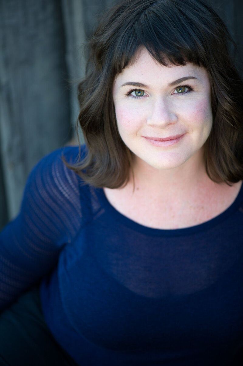 Theatrical headshot of actress Katie Adams, with shoulder length brown hair and bangs, wearing dark blue shirt.
