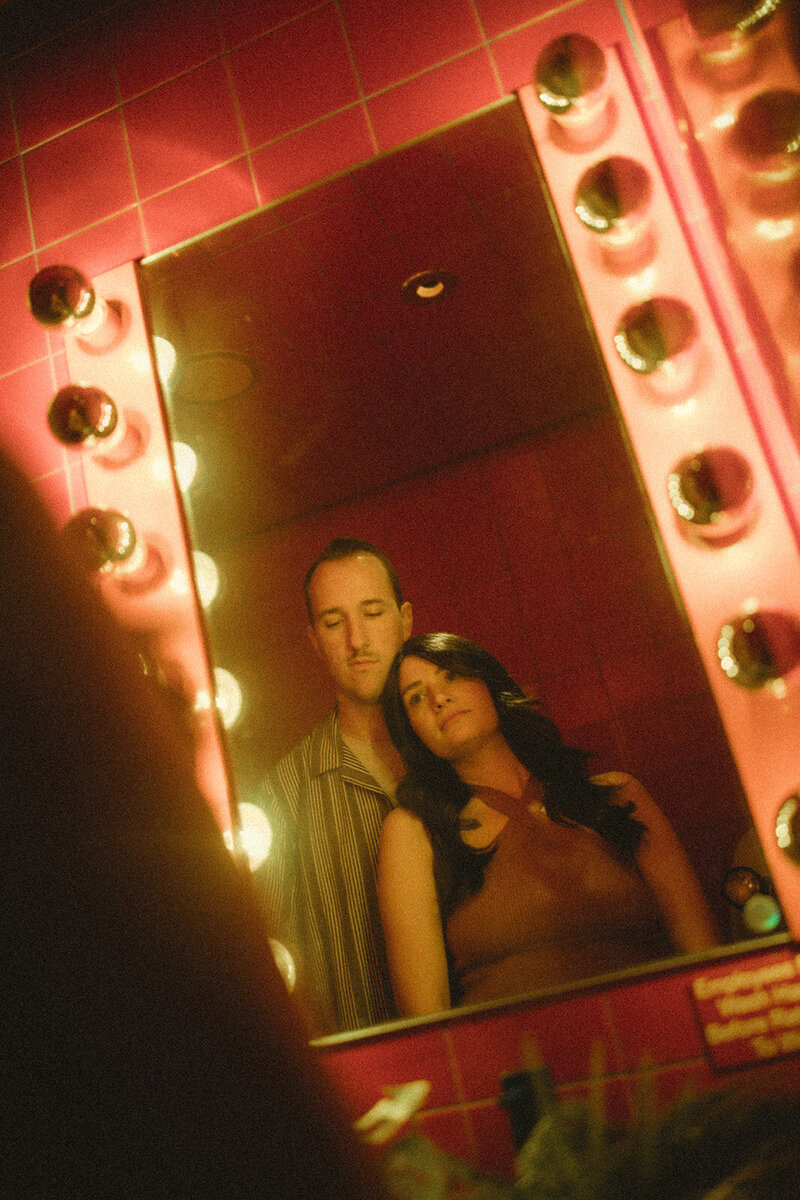A moody and intimate moment from an engagement session, featuring a couple captured in a warmly lit, retro-style mirror reflection, surrounded by vibrant pink tiles and glowing bulbs.