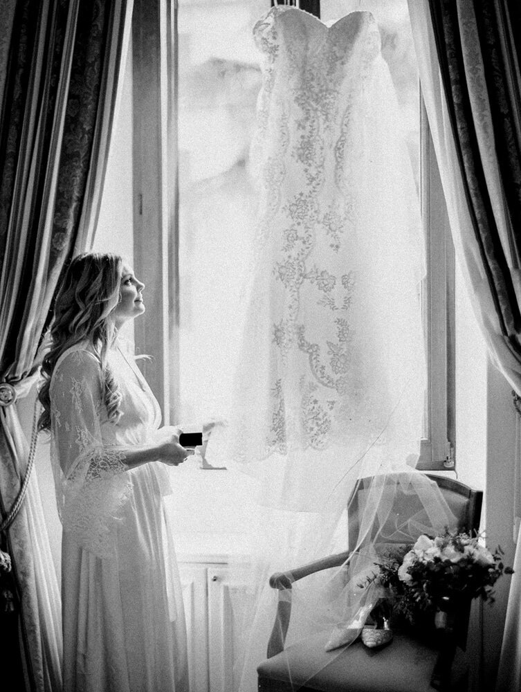 Black and white photo of a bride in a lace gown admiring her lace wedding dress that is hanging in the window.