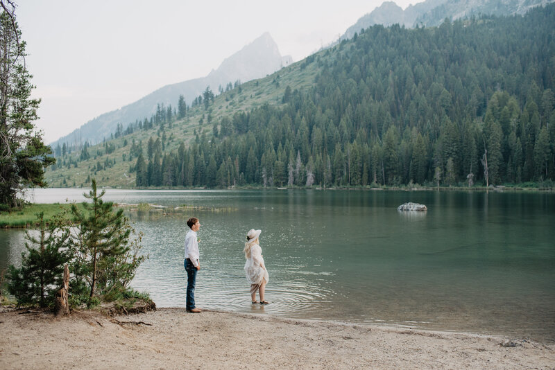 string lake wedding pictures for grand teton wedding picture with bride walking into the lake as he groom watches her from the shore with the mountains towering over the forest filled hill
