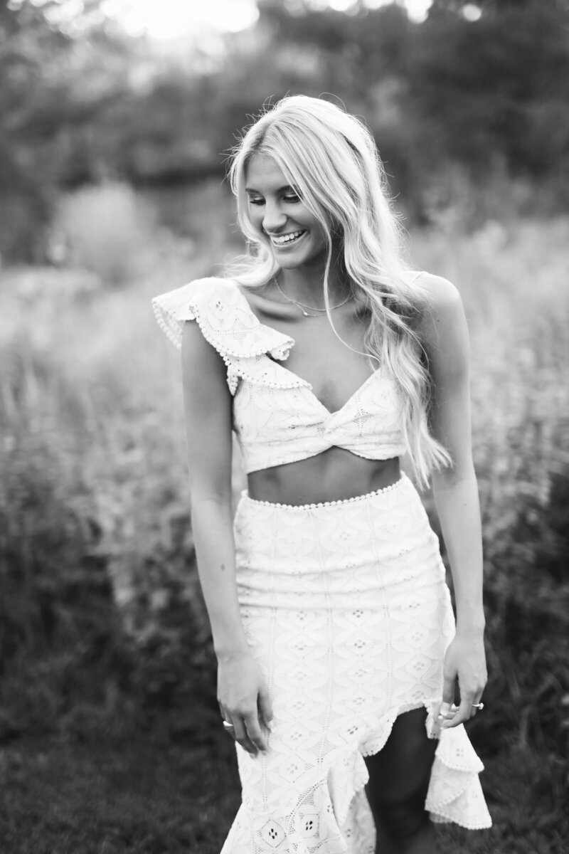 A woman with long, wavy hair walks away from the camera down a path surrounded by tall grass. She is wearing a flowing, cropped, two-piece dress. The photo, captured by a St. Louis photographer, is black and white with the sun setting in the background, casting a soft light over the scene.