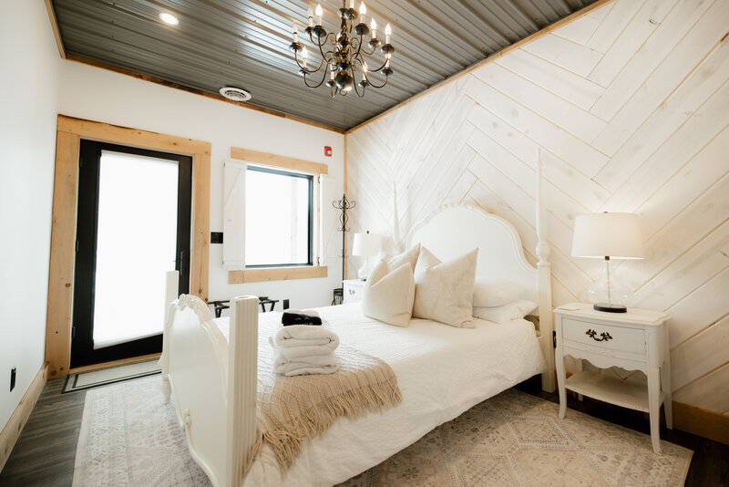 Elegant white bedroom with herringbone wood accent wall, cozy bedding, vintage nightstands, and a chandelier at Bear Ridge Destination.

