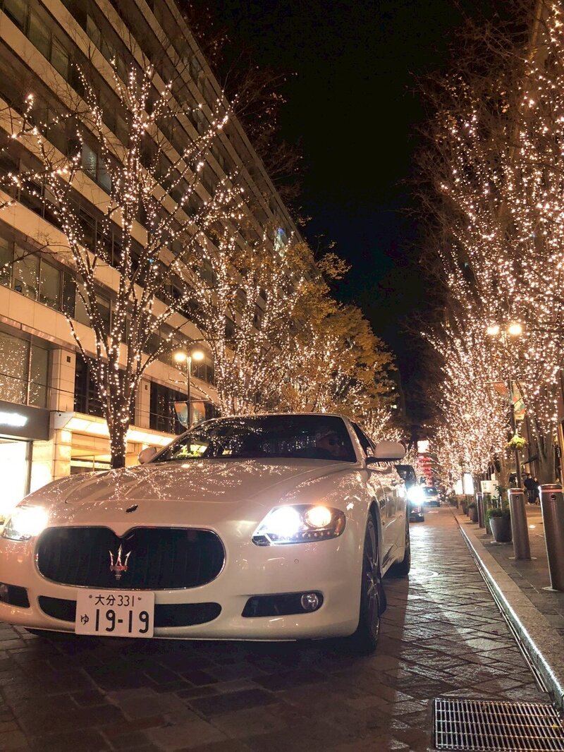 A white Maserati parked on a stylish Tokyo street lined with glowing trees and festive city lights at night.