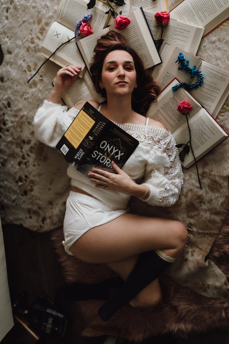 Woman lies on a soft rug surrounded by open books and roses, holding a fantasy novel during her dreamy Tampa boudoir session.