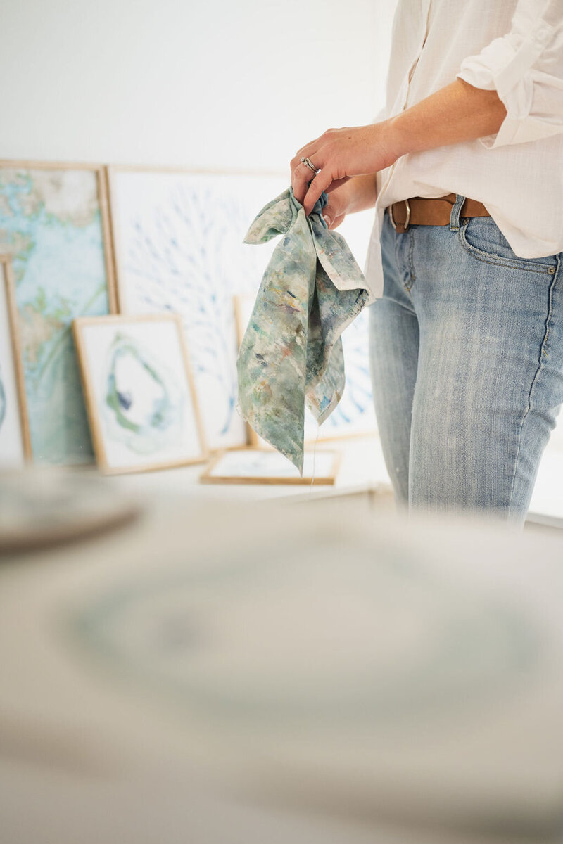 watercolor artist wearing a white button up and light jeans sitting on a wooden floor holding a tea towel with her coastal artwork displayed in the background in wilmington, north carolina